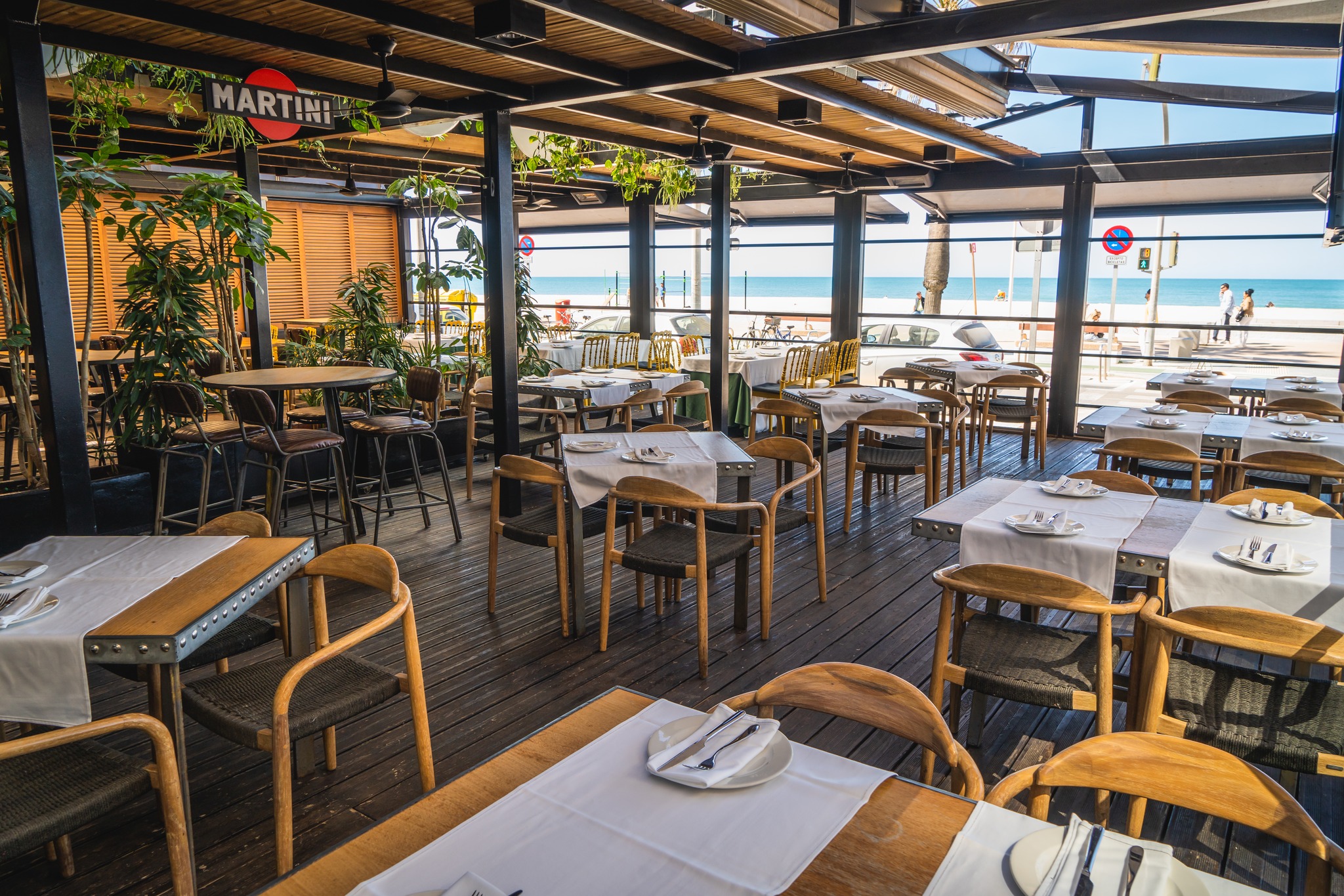 Terraza de Musalima, con mesas y sillas de madera, manteles blancos, plantas decorativas y vistas al paseo marítimo y al mar.