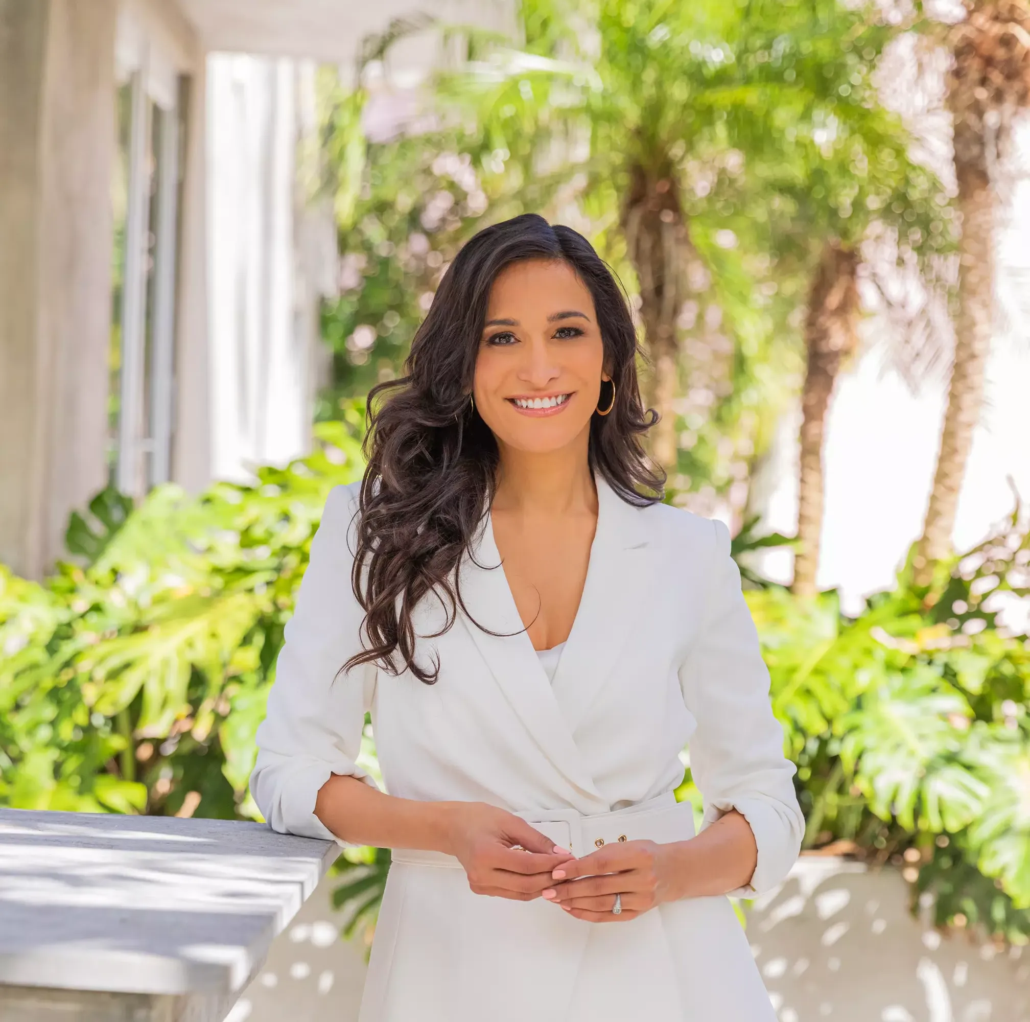 A woman in a white suit stands outdoors, smiling, with green plants and trees in the background.