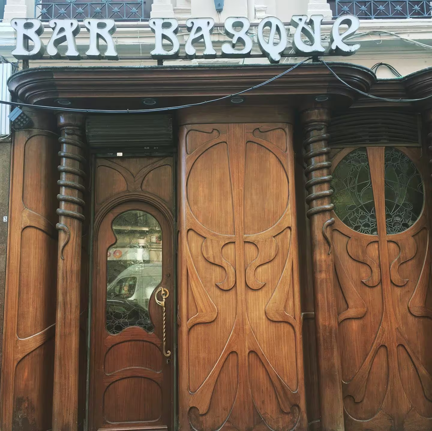 Fachada de madera de Bar Basque con diseños curvos art nouveau, puerta central acristalada y letrero en neón sobre la entrada.