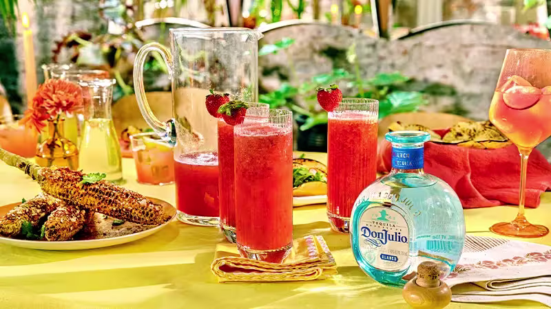 A table set with glasses of strawberry cocktails, a bottle of Don Julio tequila, grilled corn on the cob, and various dishes on a yellow tablecloth.