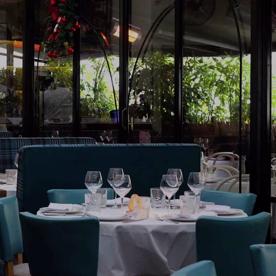 La foto muestra una mesa de comedor interior de la Taberna Los Gallos. Hay mesas con sillas tapizadas en azul y manteles blancos, con copas de vino, servilletas y cubiertos dispuestos.
