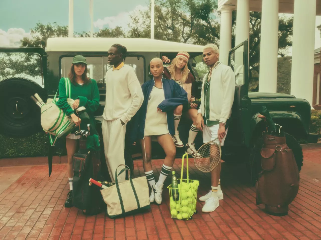 Six young adults in tennis outfits pose in front of a green SUV with tennis equipment and sports bags on a brick driveway.