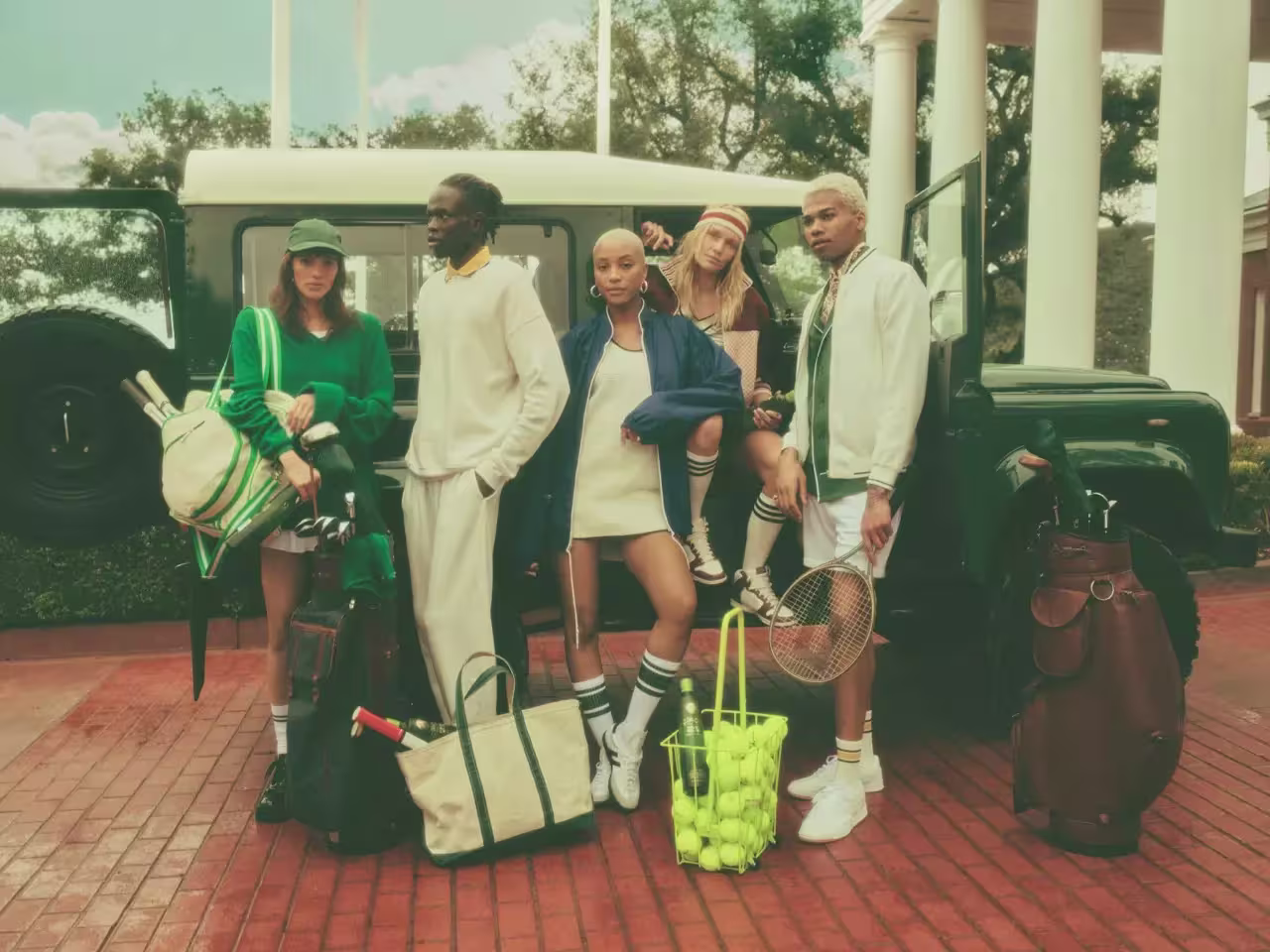 Six young adults in tennis outfits pose in front of a green SUV with tennis equipment and sports bags on a brick driveway.