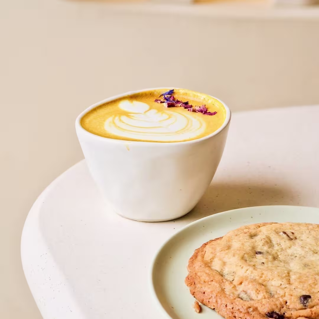 Latte dorado con arte en la espuma y pétalos secos, acompañado de una cookie con chips de chocolate sobre un plato verde pastel en una mesa de tono claro.
