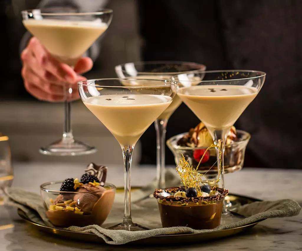 Four creamy cocktails in martini glasses are served with assorted chocolate and berry desserts on a tray, with a person reaching for one glass.