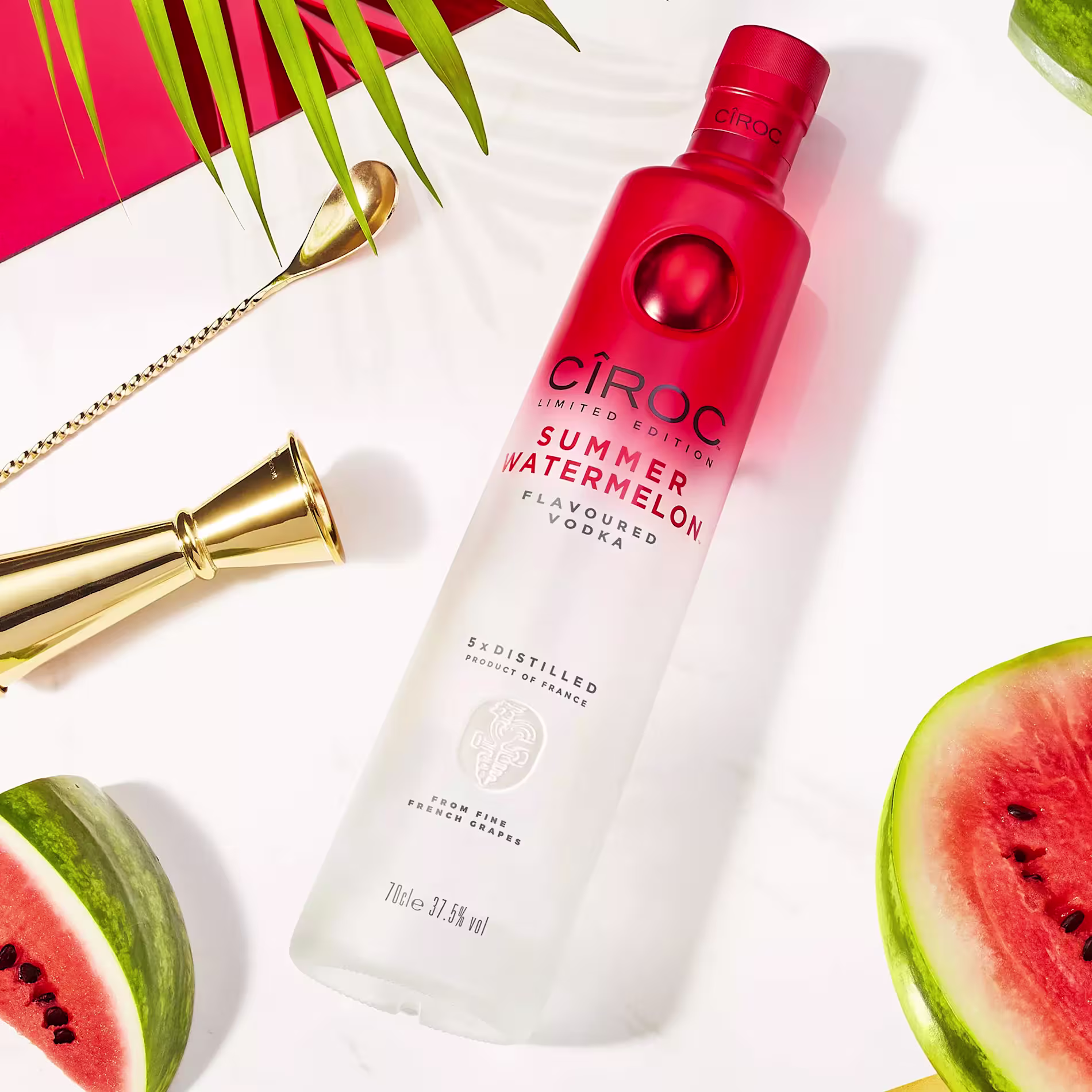 A bottle of Ciroc Summer Watermelon vodka lies on a white surface, surrounded by watermelon slices, a gold cocktail jigger, and palm leaves.