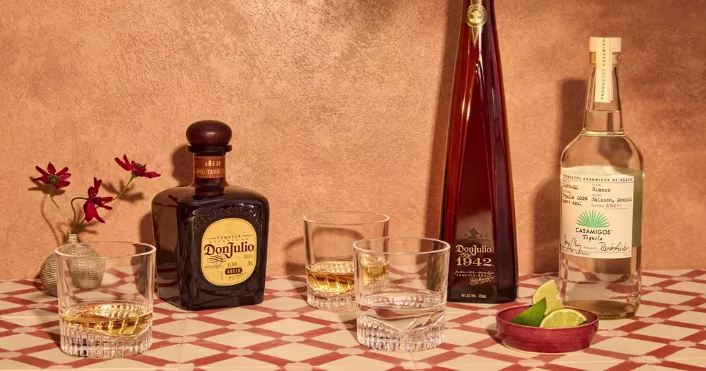 Three bottles of tequila, three glasses with drinks, and a dish with lime wedges arranged on a patterned surface with a small vase of red flowers.