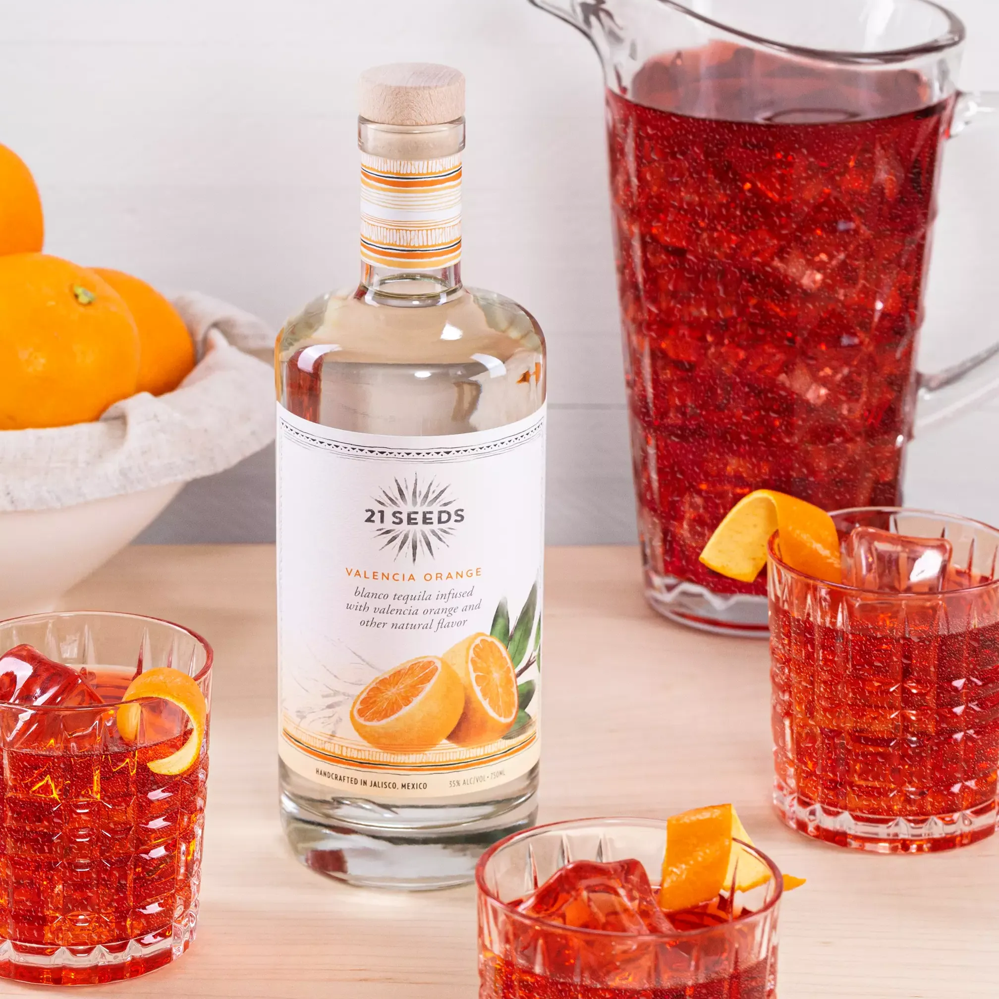 A bottle of 21Seeds Valencia Orange tequila, a pitcher, and three glasses of a red-orange cocktail with orange garnishes are arranged on a light wooden table; whole oranges are in the background.