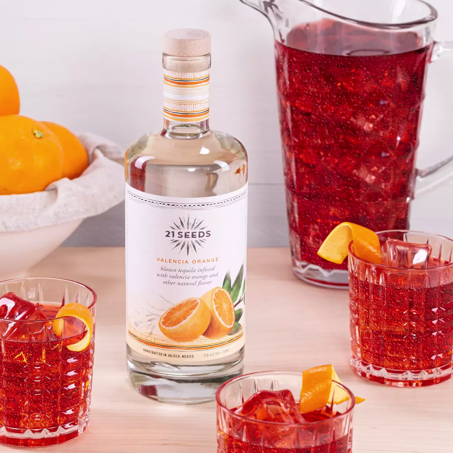 A bottle of 21Seeds Valencia Orange tequila, a pitcher, and three glasses of a red-orange cocktail with orange garnishes are arranged on a light wooden table; whole oranges are in the background.
