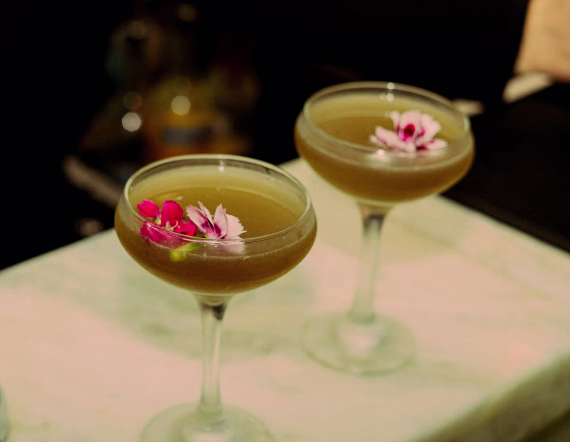 Two coupe glasses filled with brown cocktails, each garnished with pink and white edible flowers, sit on a marble countertop.