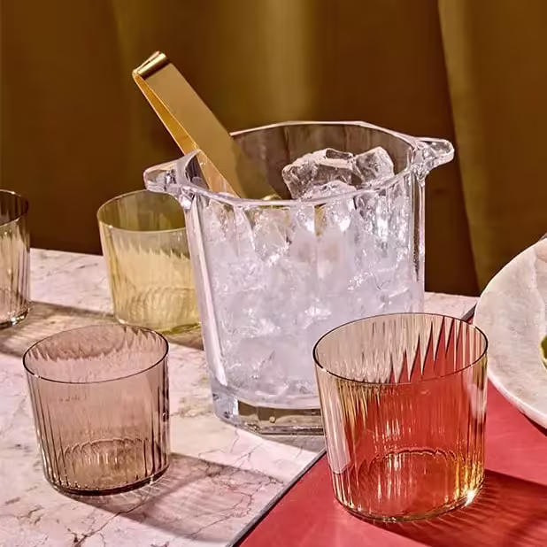 A glass ice bucket with a gold tong and ice cubes is placed on a table with four colored glass tumblers and part of a white plate.