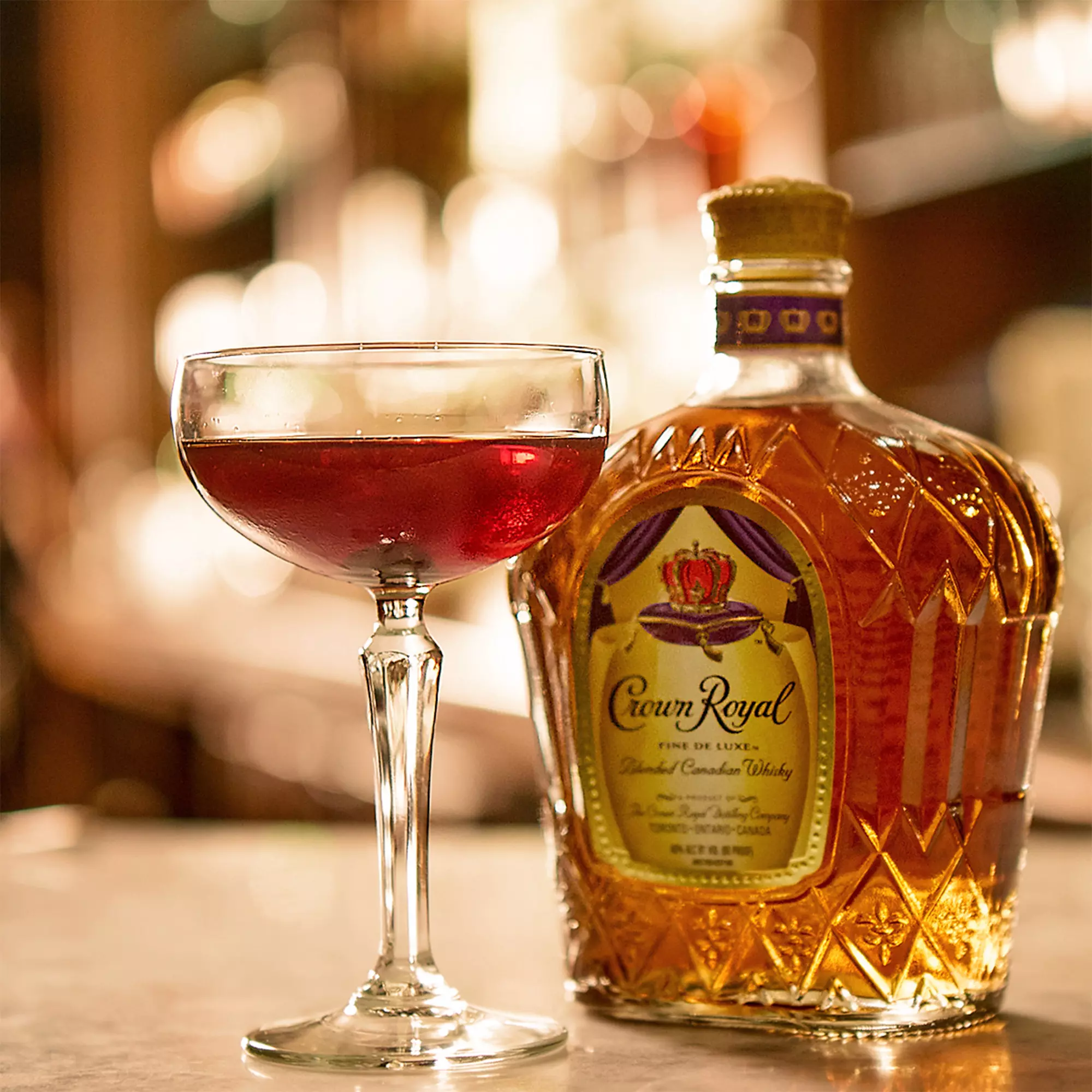 A glass of red cocktail sits next to a bottle of Crown Royal Canadian whisky on a bar counter.