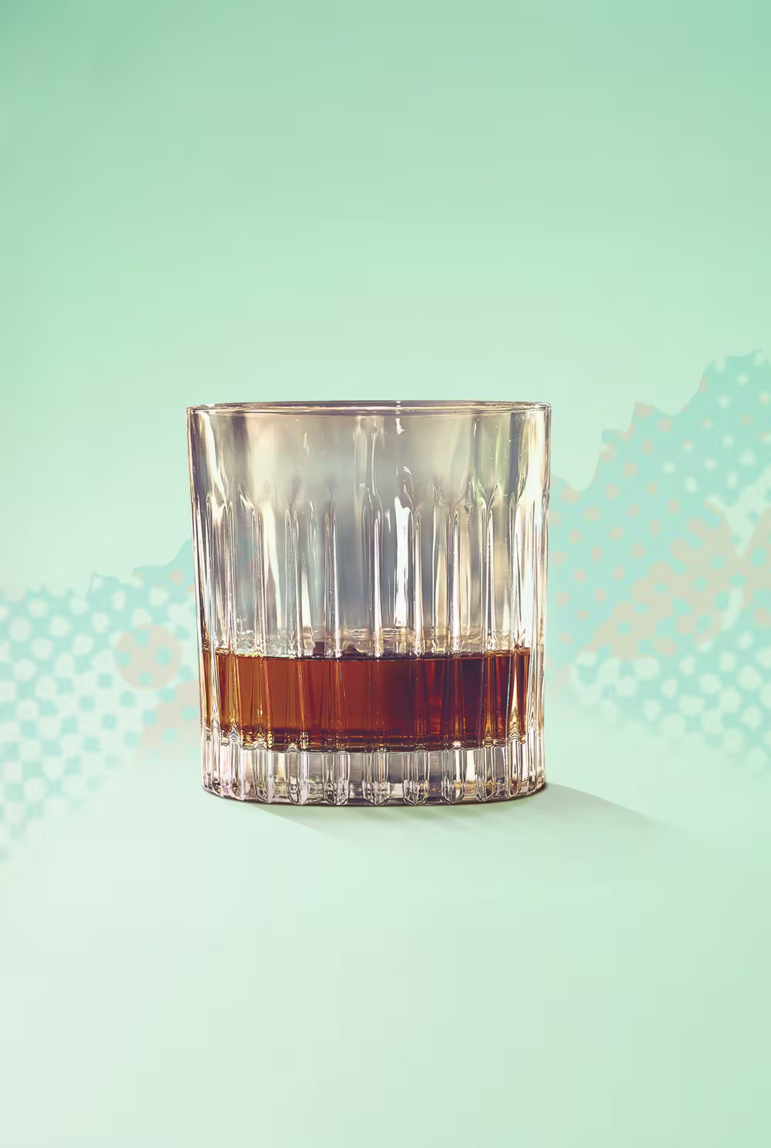 A glass tumbler with a small amount of brown liquid, likely whiskey or bourbon, sits on a light green surface with a patterned background.