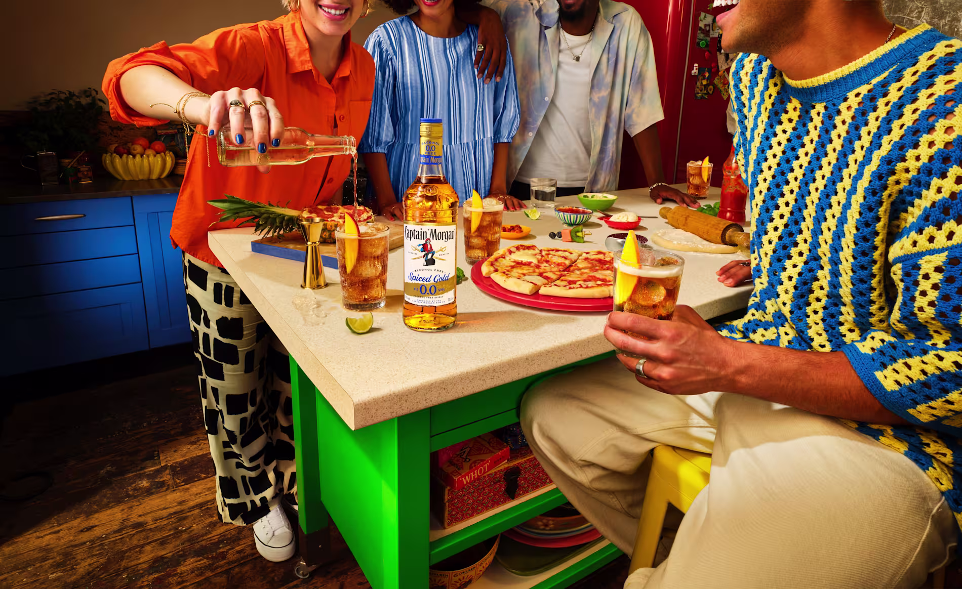 People enjoying Captain Morgan Spiced Gold 0.0% cocktails with pizza at a colorful kitchen counter