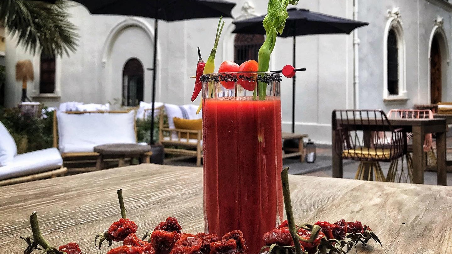 Cóctel rojo con apio, tomates y chile servido en la terraza de Cooktl by La Salita, sobre una mesa rústica con decoración de pimientos secos y carta negra.