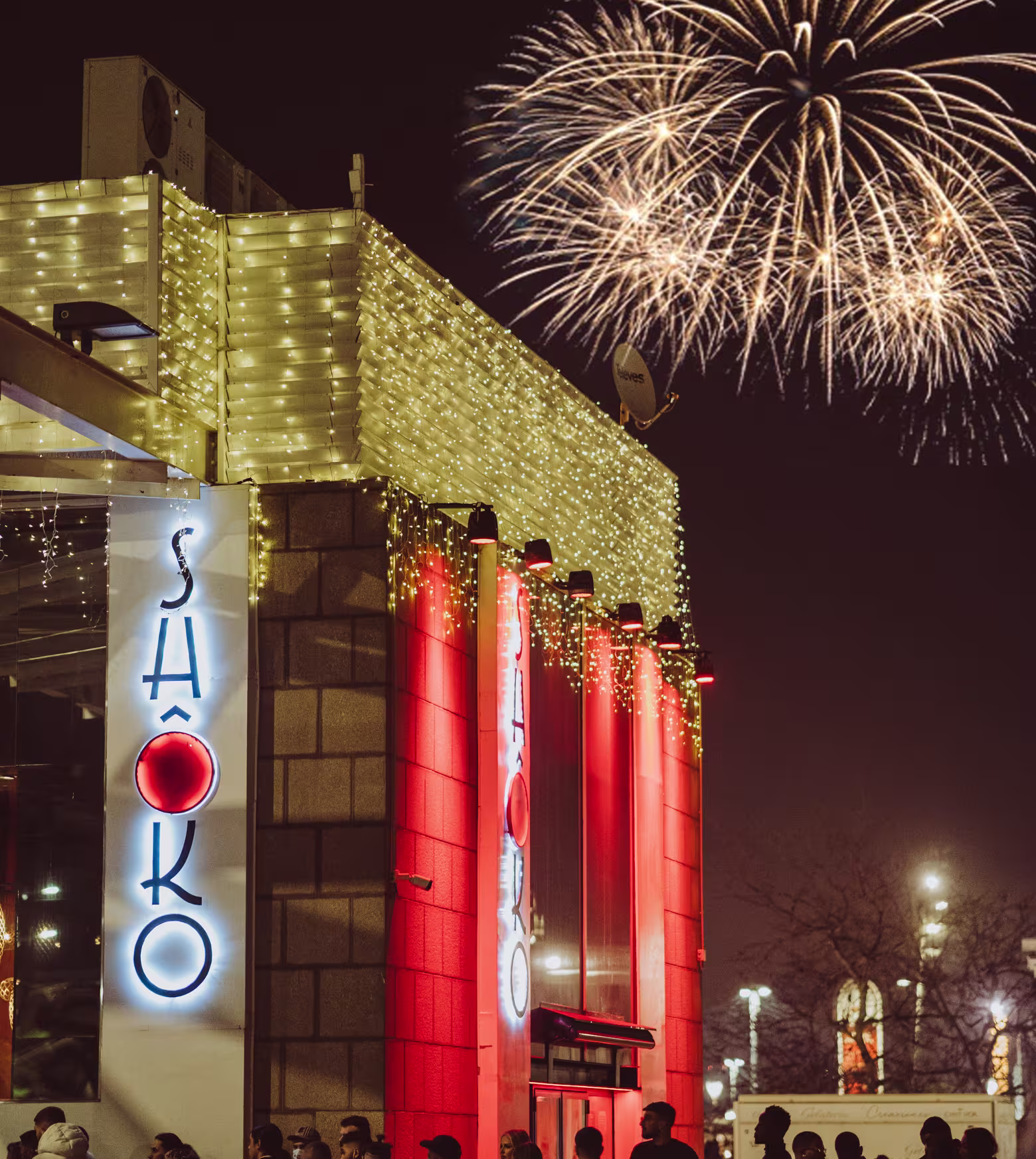Fachada de Shôko Barcelona Club iluminada con luces festivas, detalles rojos vibrantes y fuegos artificiales de fondo.