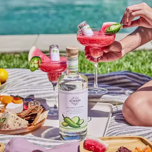 A bottle of 21 Seeds Cucumber Jalapeño tequila sits on a picnic blanket next to snacks, with two watermelon margaritas held by people near a pool.
