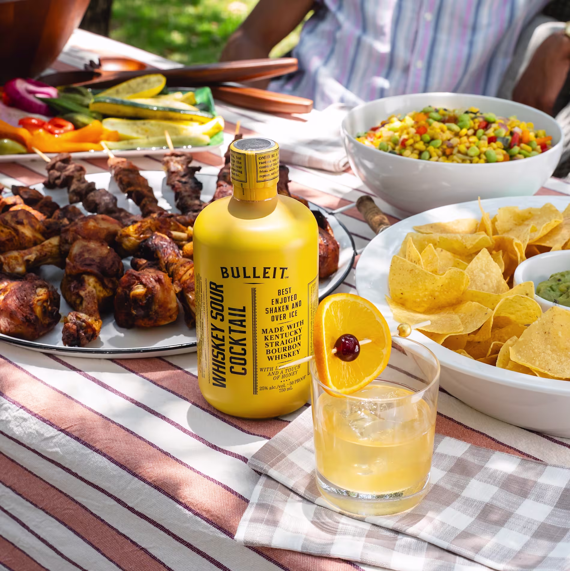 A picnic table with grilled meats, vegetables, chips with dip, corn salad, and a Bulleit Whiskey Sour cocktail bottle next to a glass with a cherry and orange slice garnish.