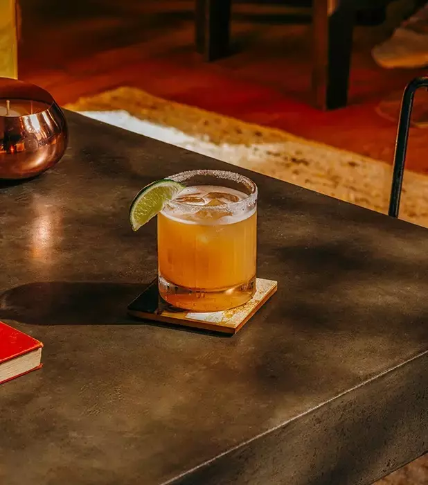 A cocktail with a lime wedge sits on a square coaster on a dark table in a warmly lit room.
