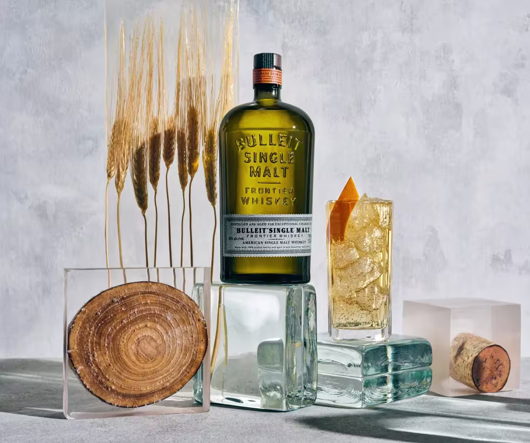 Botella de whisky de la amrca Bulleit, sobre un fondo gris y unos cubos de hielo decorados y un cocktail al lado naranja.