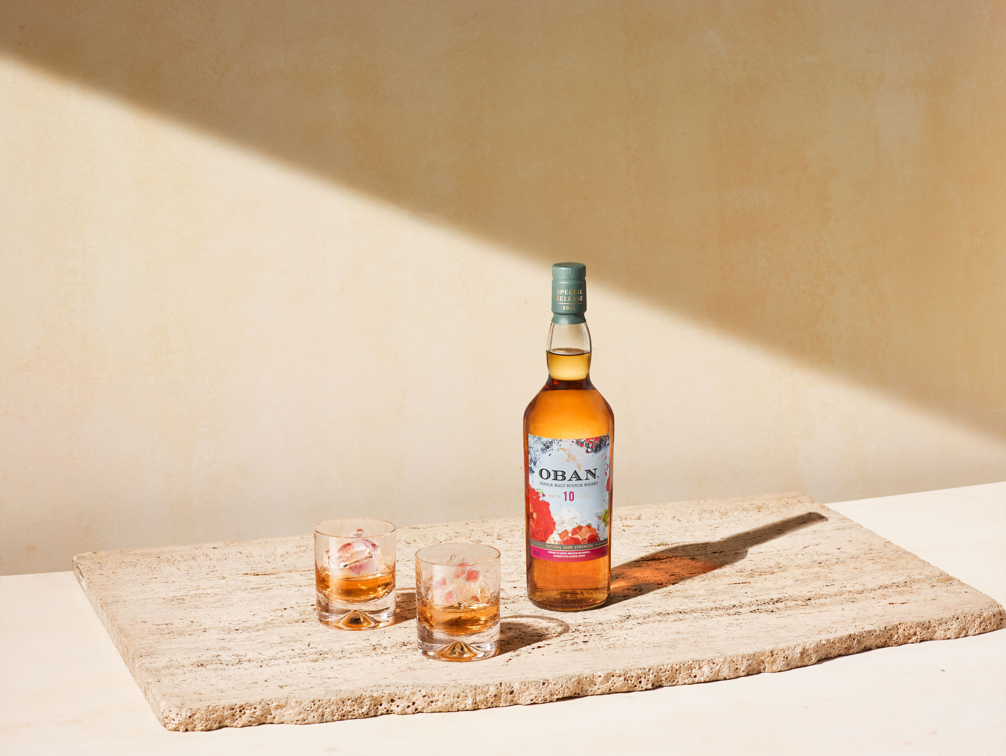 A bottle of Oban 10 whisky stands on a stone slab next to two glasses filled with whisky and ice, against a beige background with a diagonal shadow.