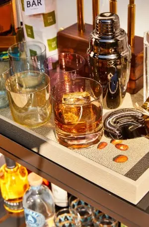 A bar cart with assorted glasses, a cocktail shaker, bottles, a tray with ice-filled whiskey glass, almonds, and a corkscrew.