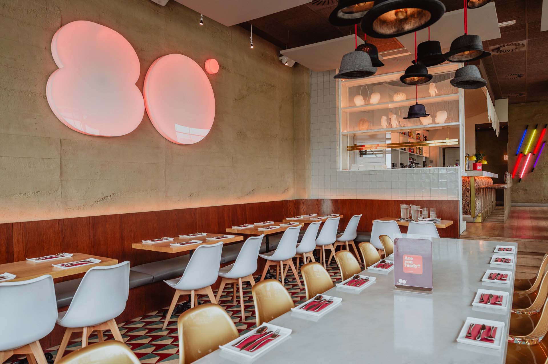 Interior de Ochenta Grados Las Tablas con decoración moderna, logo retroiluminado en rosa, suelo geométrico y lámparas en forma de sombrero sobre mesas largas.