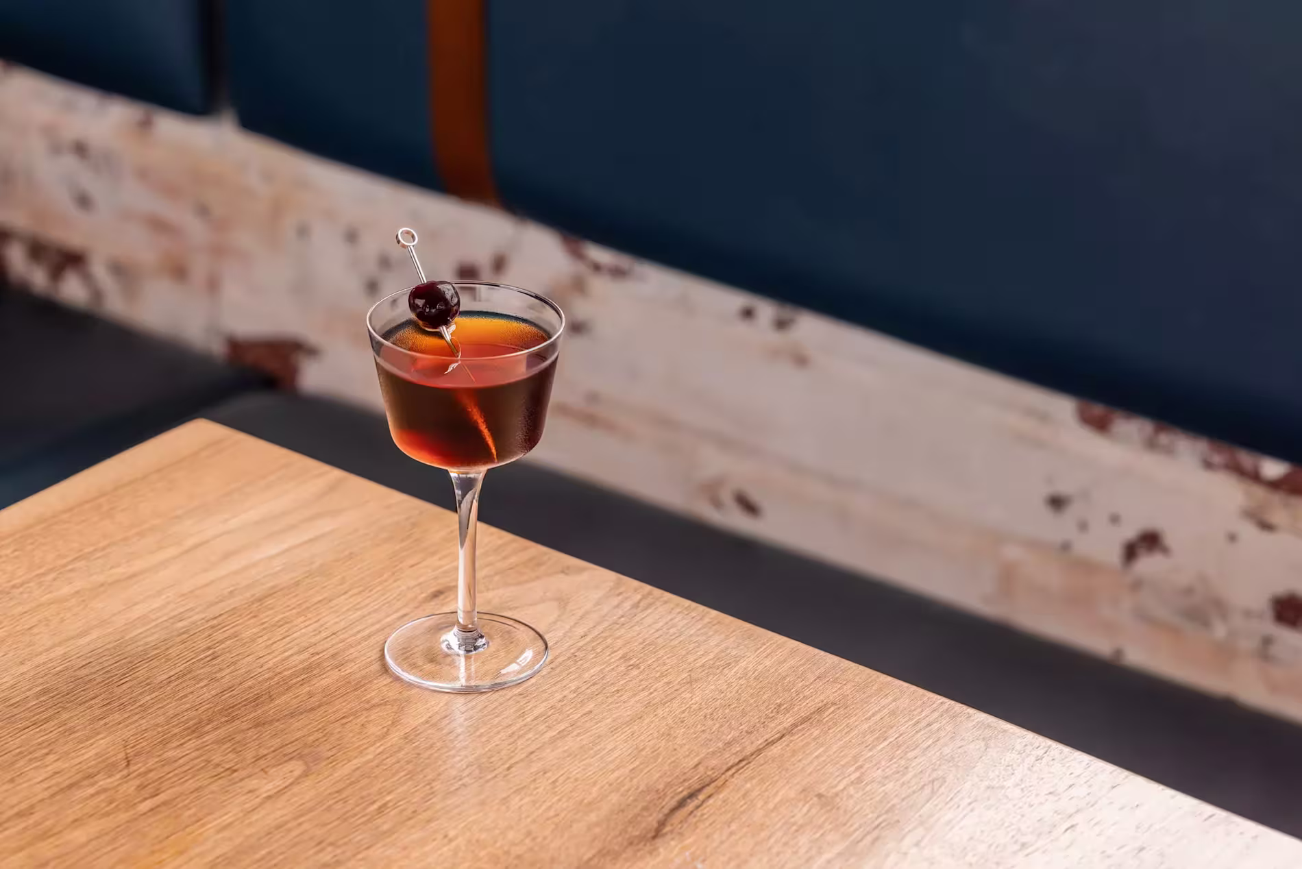 A cocktail in a stemmed glass, garnished with a cherry on a pick, sits on a wooden table with a blue cushioned bench in the background.