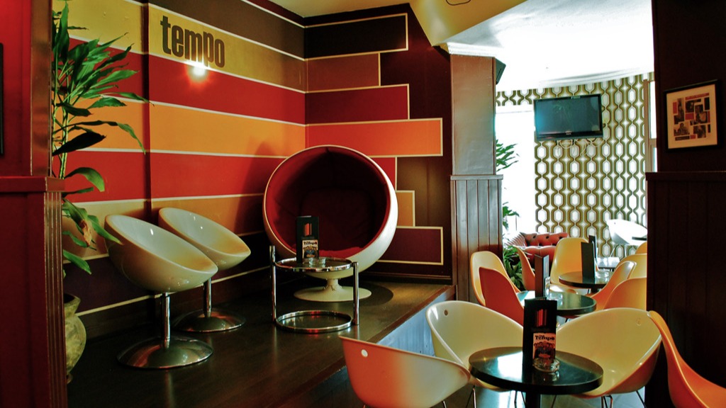 Paredes de tonos cálidos, sillas futuristas y una icónica cápsula roja crean un ambiente retro en Tempo Club, con luz suave y decoración geométrica.