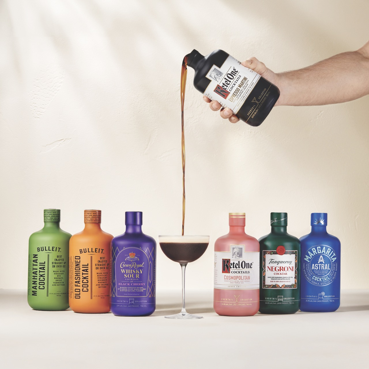 Six colorful cocktail bottles are lined up side by side, with a hand pouring a dark drink from one bottle into a cocktail glass in the center.