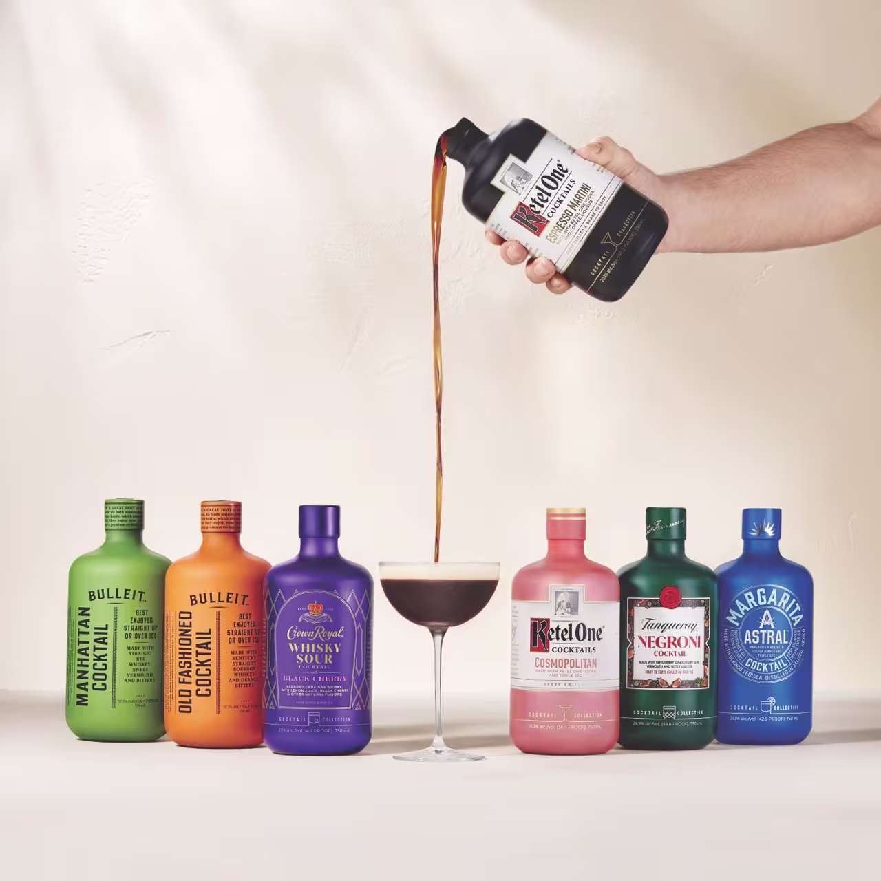 Six colorful cocktail bottles are lined up side by side, with a hand pouring a dark drink from one bottle into a cocktail glass in the center.