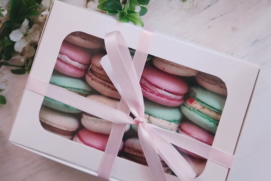 A box of assorted macarons in pastel colors is tied with a light pink ribbon and placed on a light-colored surface with some small flowers nearby.
