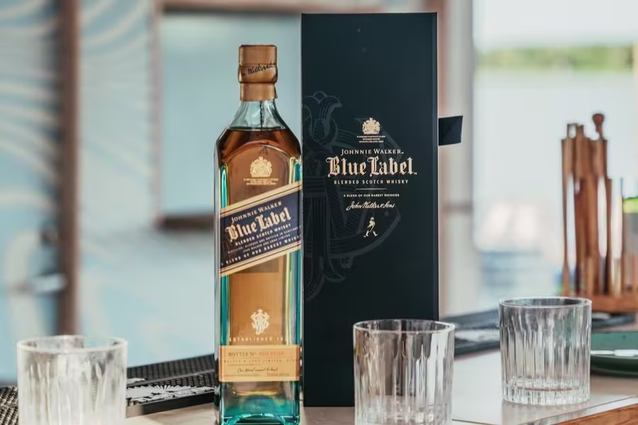 A bottle of Johnnie Walker Blue Label whisky stands next to its box on a table with three empty glass tumblers.