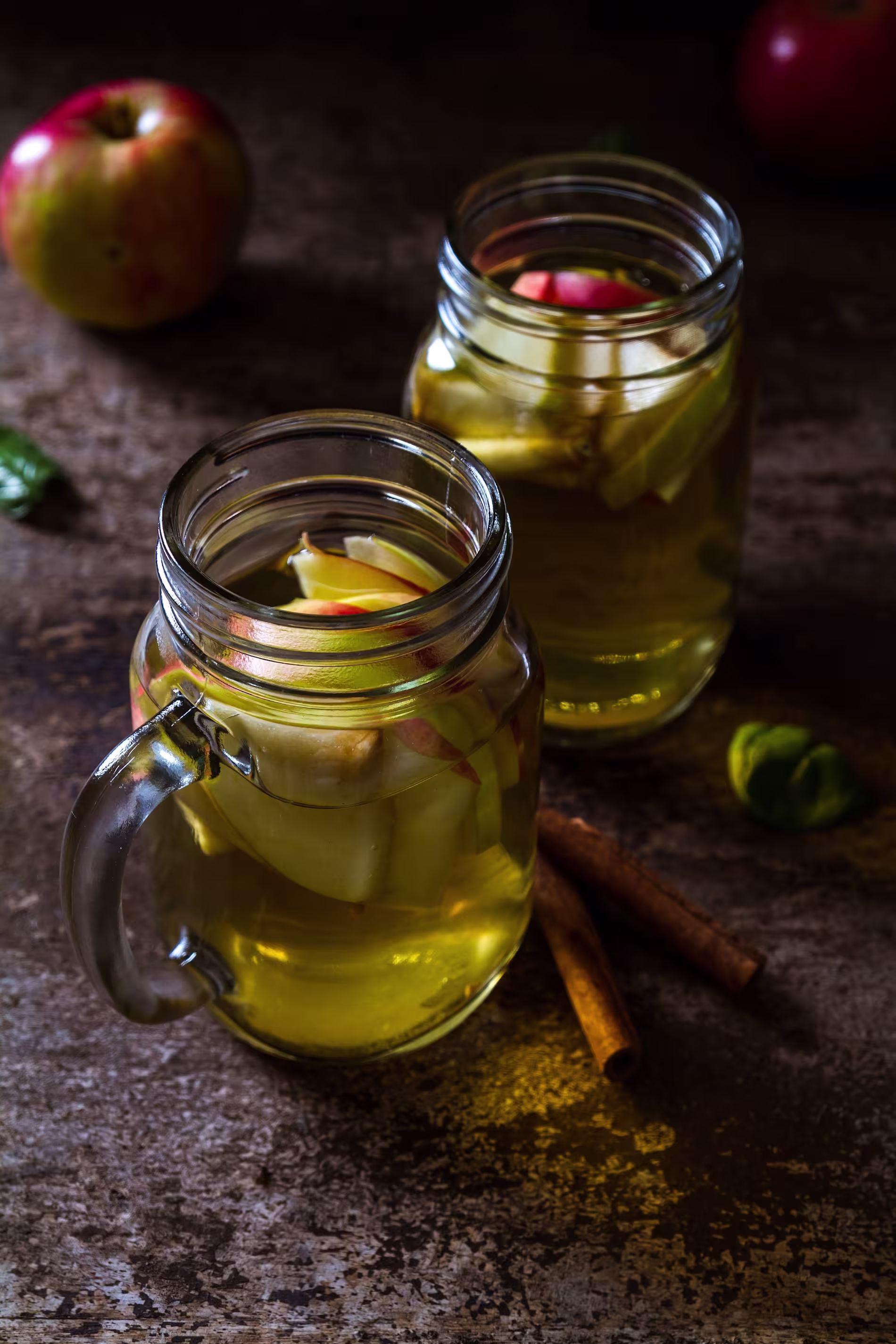 Two mason jar mugs filled with apple slices and a yellowish liquid sit on a rustic surface, with cinnamon sticks, an apple, and green leaves nearby.