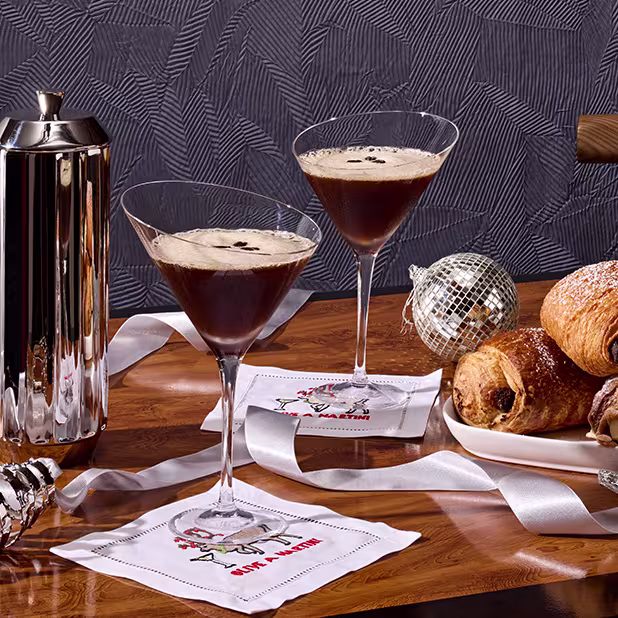 Two espresso martinis on cocktail napkins, a French press, pastries on a plate, silver ribbon, and holiday ornaments on a wooden table.