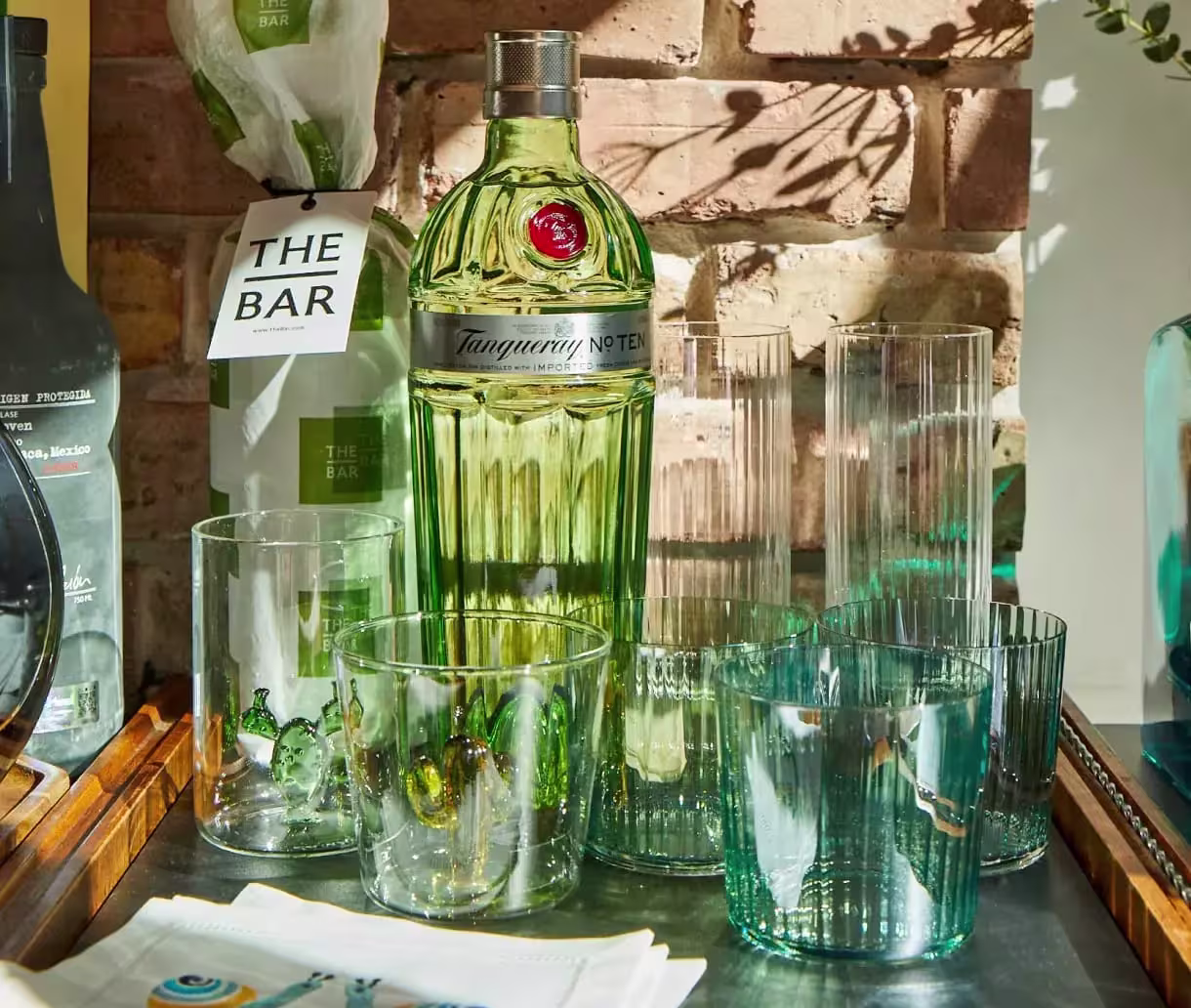 A bottle of Tanqueray gin is placed on a table surrounded by various clear drinking glasses, with a brick wall and gift items in the background.