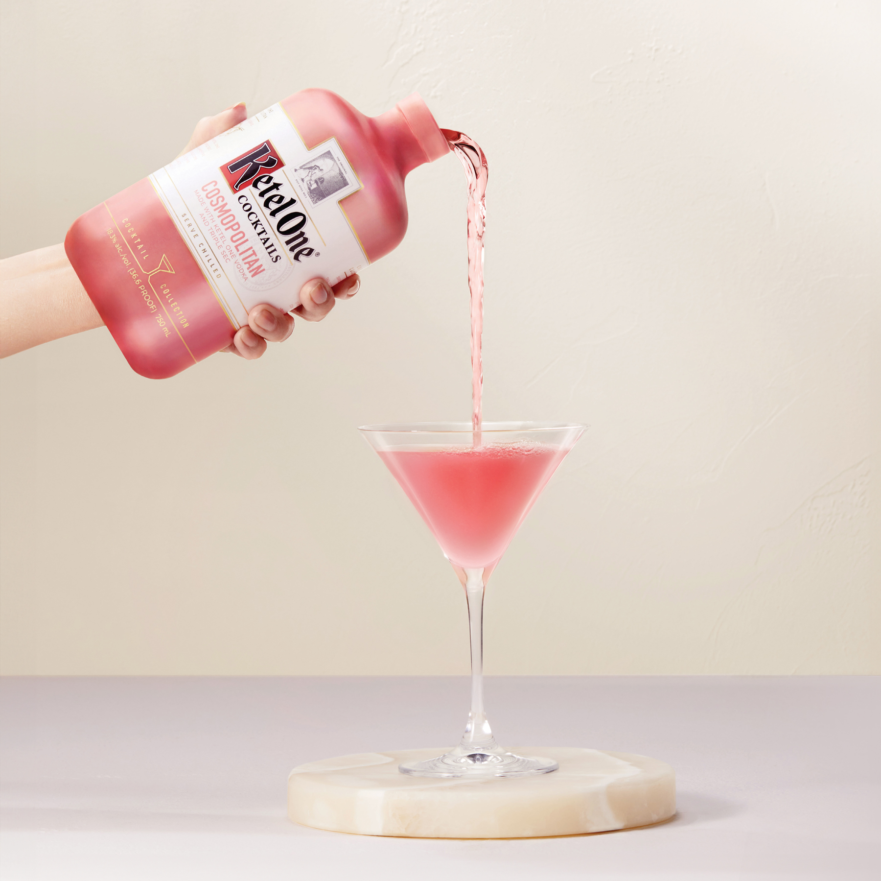 A hand pours pink RealOne Cosmopolitan Easy Pour Cocktail Mix from a bottle into a martini glass on a round marble coaster.