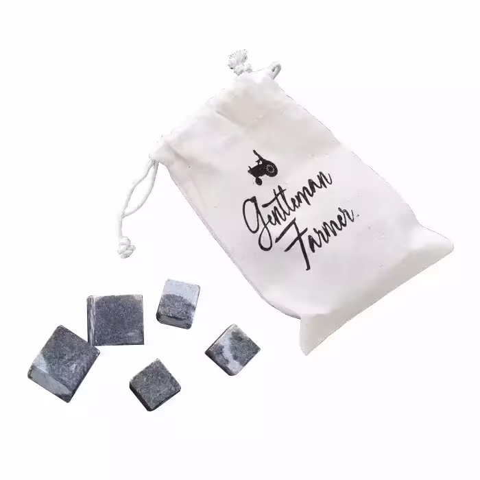 A small cloth pouch labeled “Gentleman Farmer” next to five gray whiskey stones arranged on a white surface.