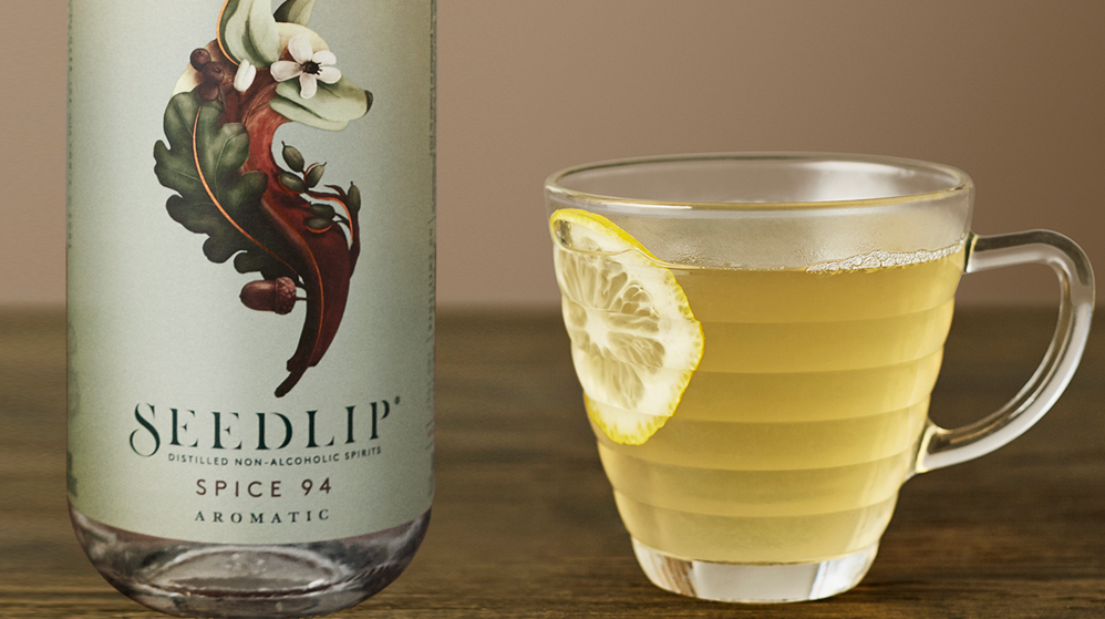 A bottle of Seedlip Spice 94 non-alcoholic spirit is next to a glass mug containing a yellow drink garnished with a lemon slice.