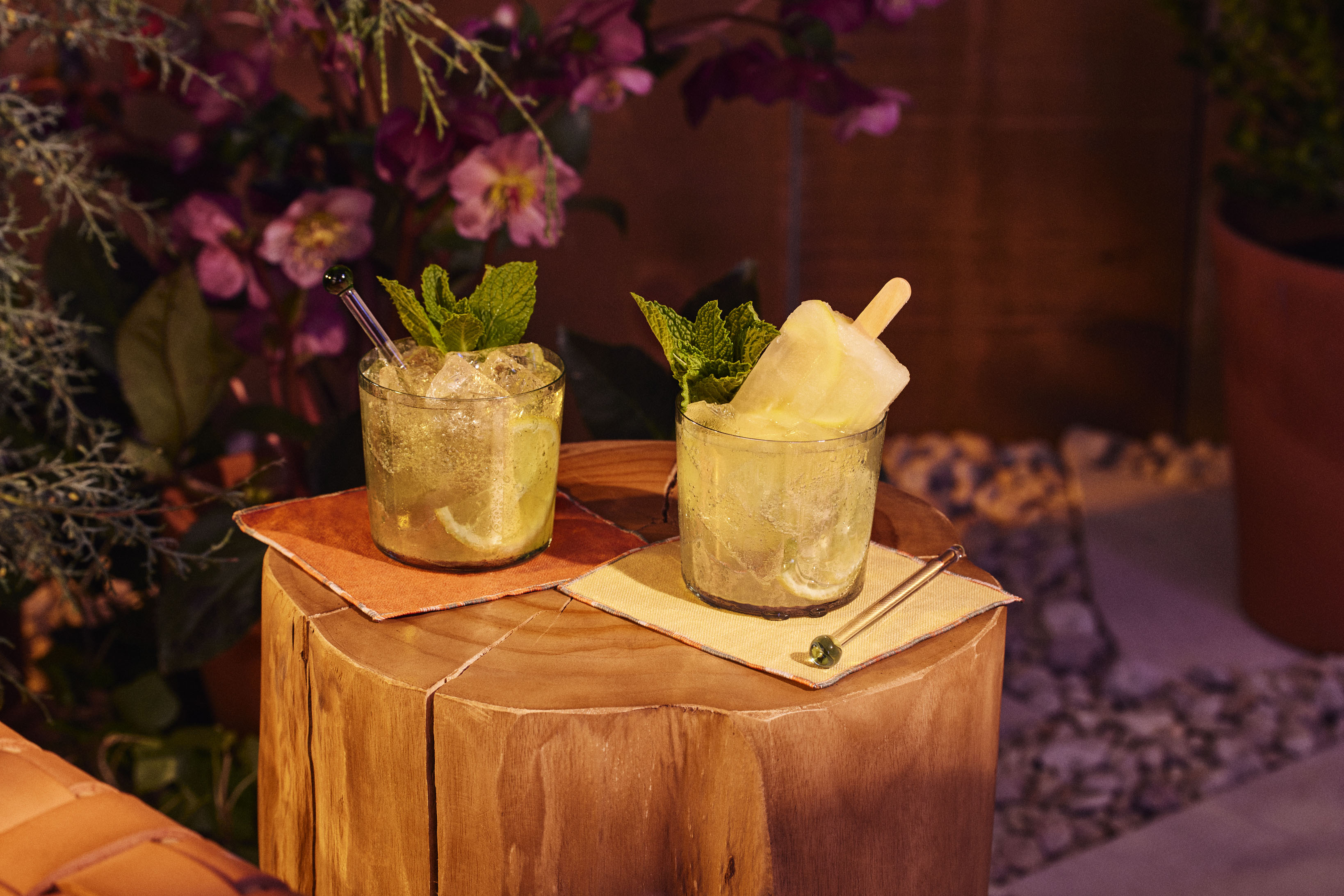 Two cocktails garnished with mint leaves, one with a popsicle, are placed on colored napkins atop a wooden stump, with flowers and plants in the background.