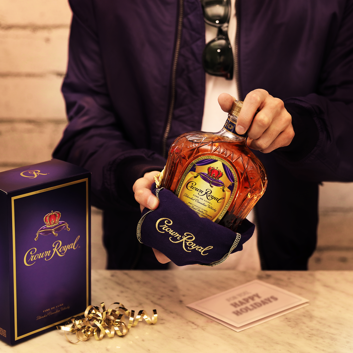 A person holds a bottle of Crown Royal whisky in a purple velvet pouch next to its box, a gold ribbon, and a card on a marble counter.