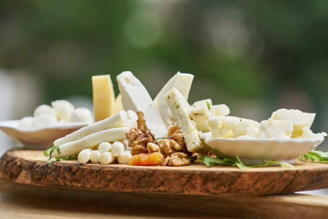 A wooden platter with assorted cheeses, walnuts, dried apricots, and two small dishes containing round and cubed cheese pieces, set outdoors.