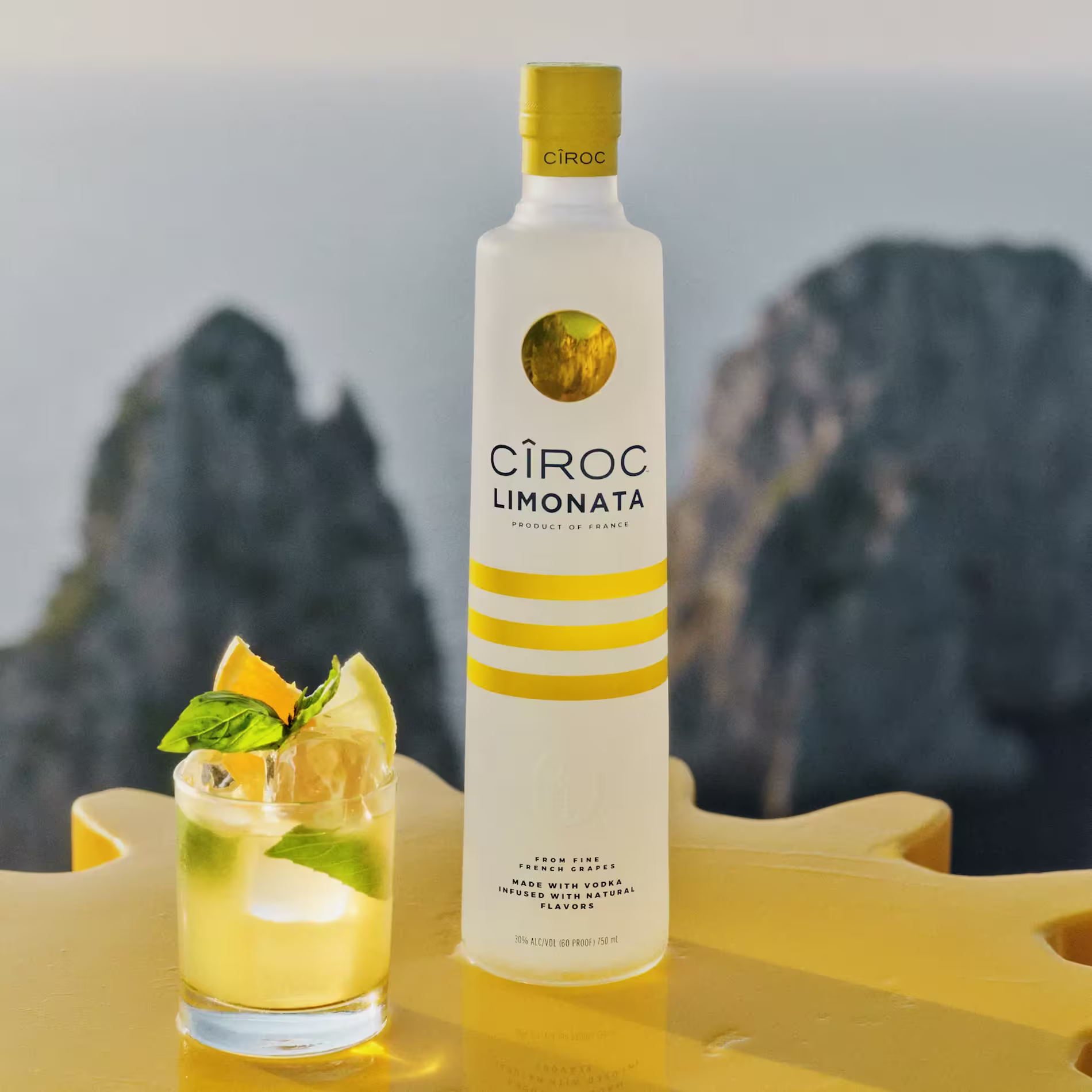 A bottle of Ciroc Limonata vodka stands next to a cocktail garnished with citrus slices and mint, set on a yellow surface with a blurred natural background.