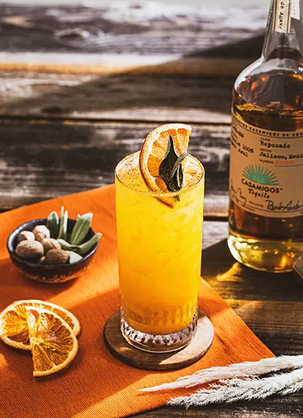 A tall glass of orange cocktail garnished with a dried orange slice and herb sits on an orange napkin beside a bottle of Casamigos tequila and dried orange slices.