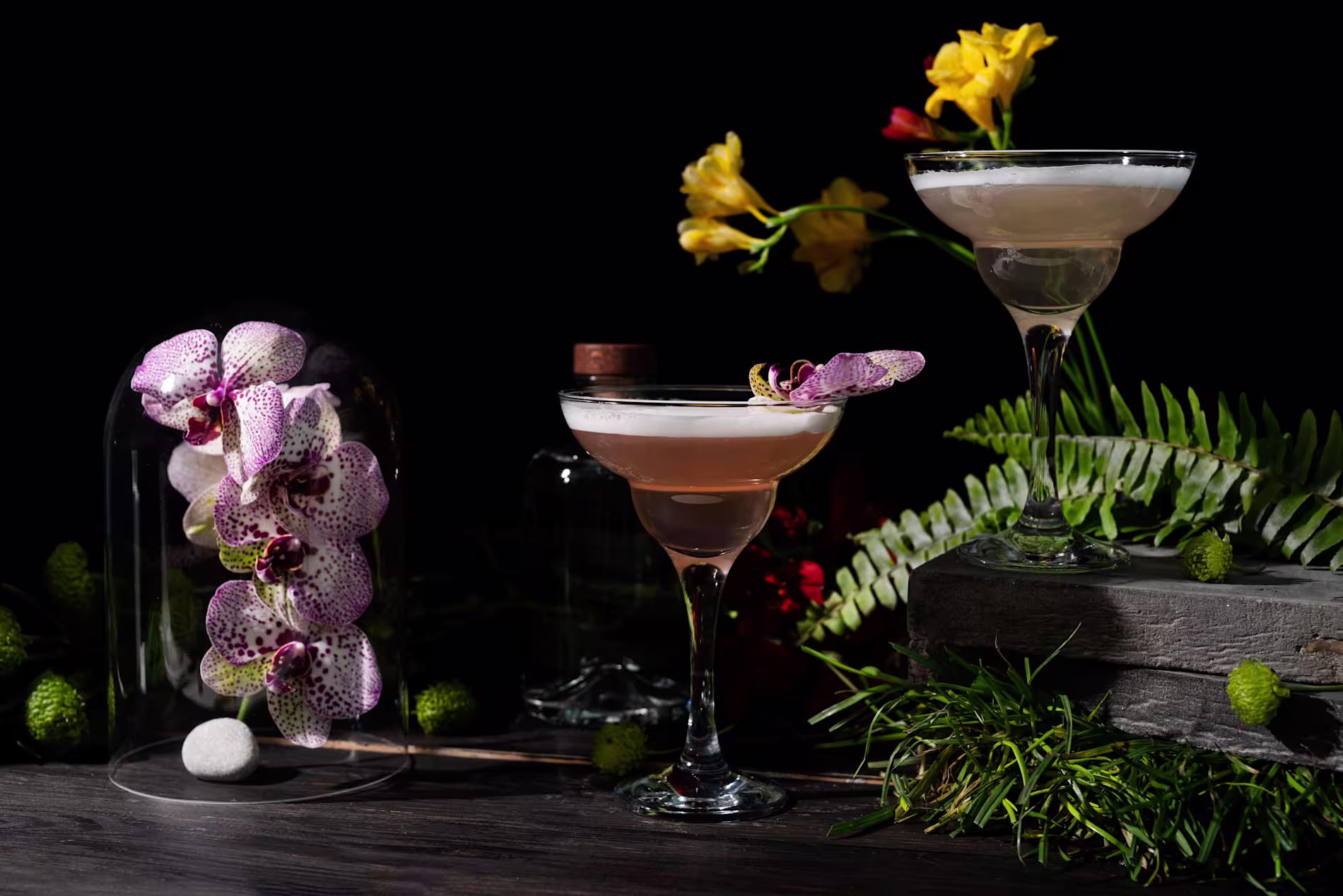 Two elegant cocktails garnished with orchids and ferns sit on a wooden surface, surrounded by flowers, greenery, a small bottle, and a glass dome containing an orchid blossom.
