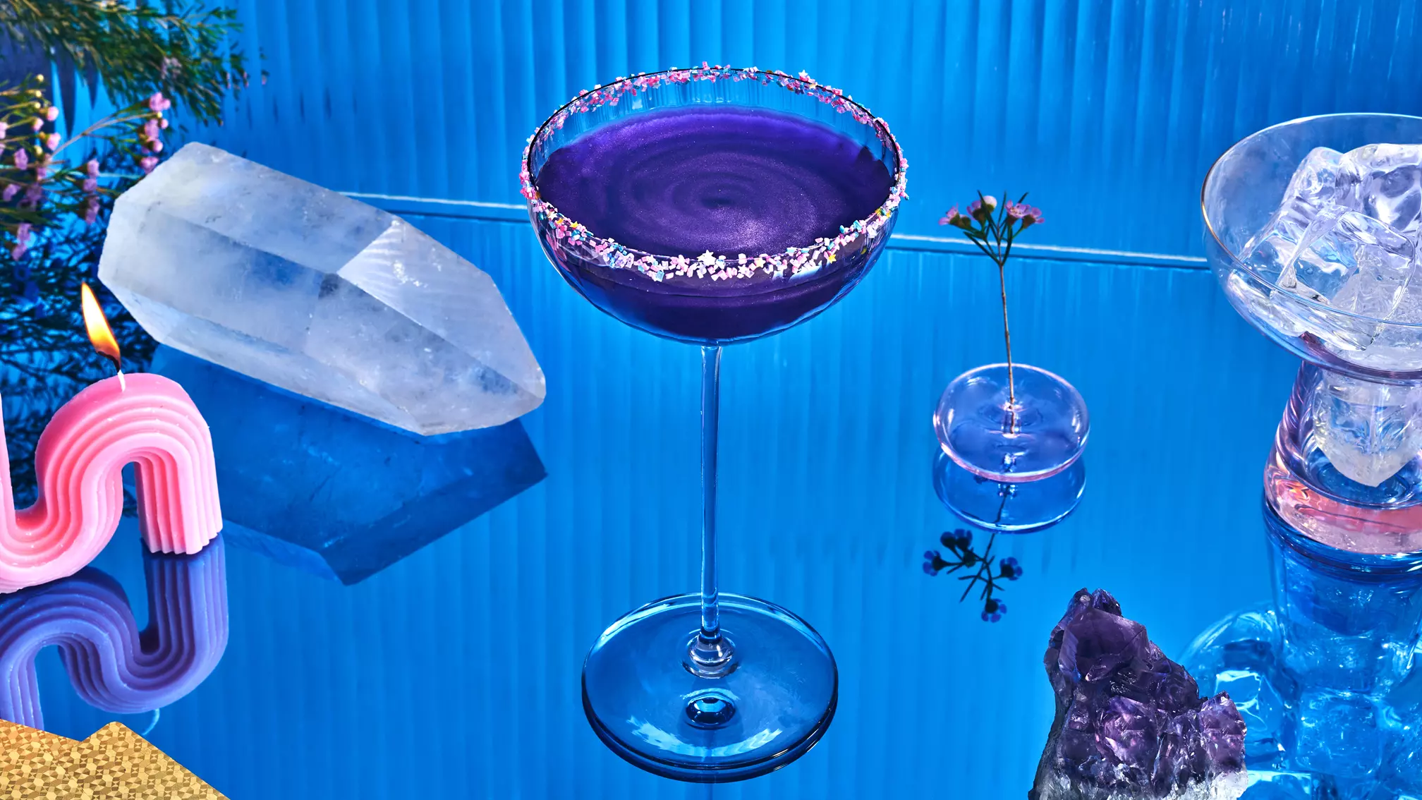 A blue cocktail in a coupe glass with a sugared rim sits on a reflective blue surface, surrounded by crystals, ice, a pink candle, and a small flower in a vase.