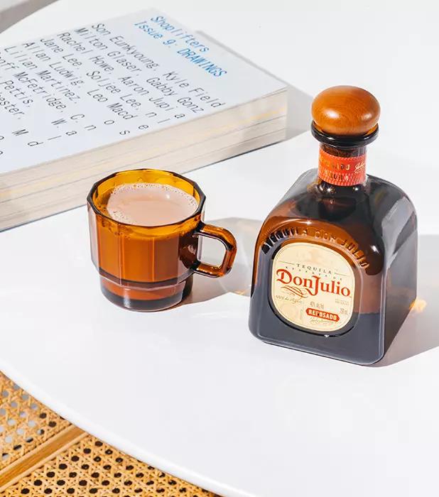 A bottle of Don Julio tequila and a brown glass cup with a creamy beverage sit on a white table next to an open book.