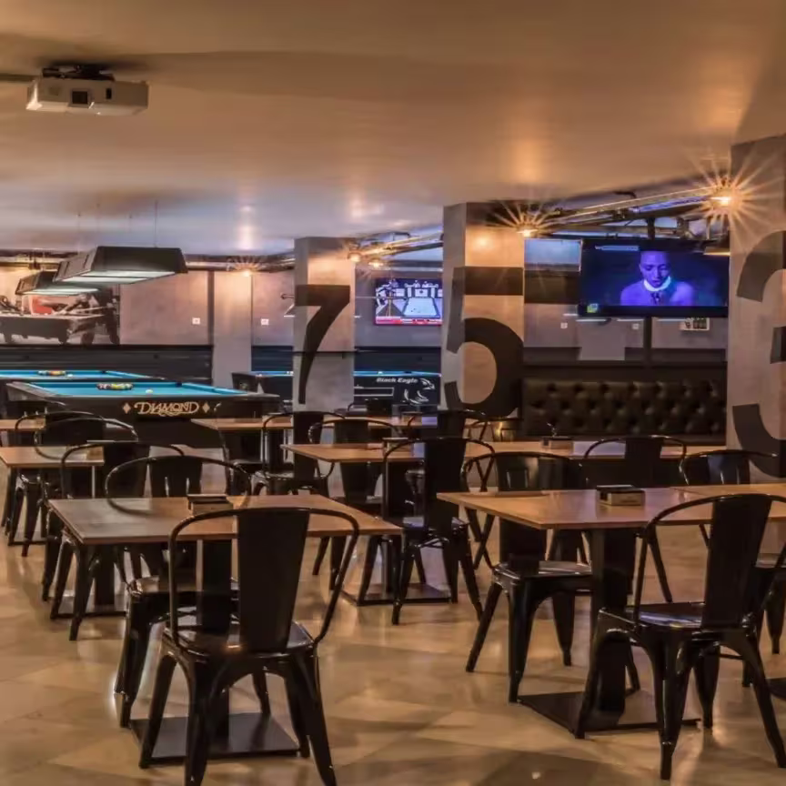 La imagen muestra el interior de La Pecera del Pool, un local amplio y moderno con mesas y sillas de estilo industrial. Destacan las mesas de billar en el fondo y pantallas para ver deportes, creando un ambiente relajado para socializar.