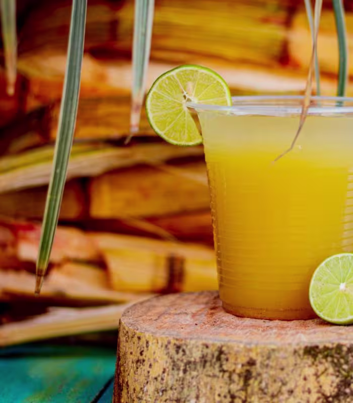 A plastic cup filled with yellow juice, garnished with two lime slices, sits on a wooden surface with sugarcane stalks in the background.