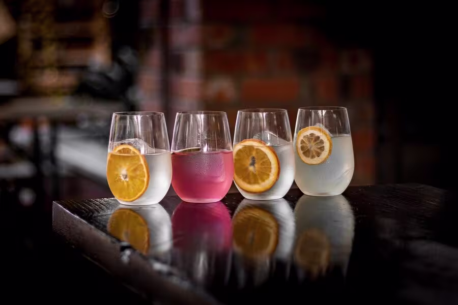 Four stemless glasses with drinks, each garnished with a citrus slice, are arranged in a row on a dark wooden surface with a blurred brick wall background.